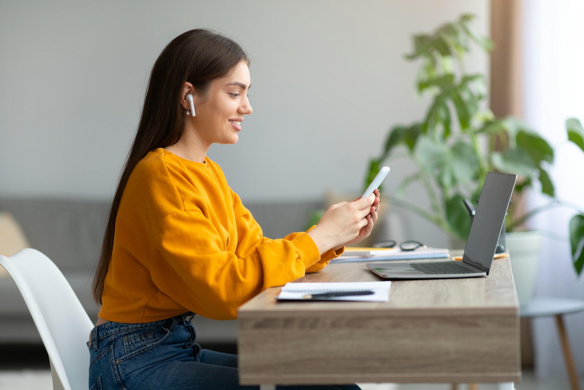 Happy young woman in earphones sitting at desk with laptop, using cell phone at home, blank space